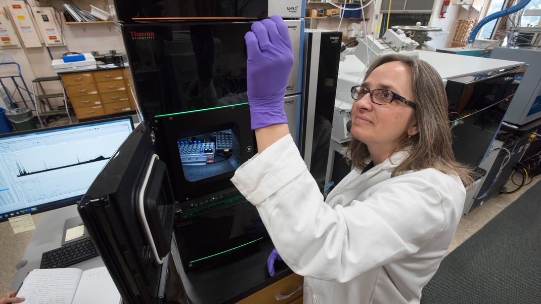 A women wearing a lab coat, gloves, and a pair of glasses holds up a sample of something. Behind her are tall, microwave-like machines and a computer monitor.