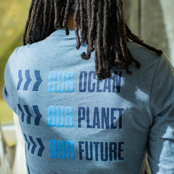 The back of a T-shirt with text reading "Our Ocean, Our Planet, Our Future," with arrow icons. The shirt is worn by a woman with long dark hair.