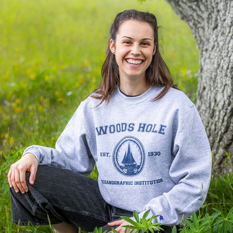 A girl kneels in the grass by a tree trunk. She's wearing a crew neck sweatshirt. The sweatshirt reads "Woods Hole," followed by the institutional seal sandwiched by "EST" and "1930," and "Oceanographic Institution."