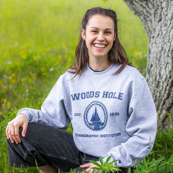 A girl kneels in the grass by a tree trunk. She's wearing a crew neck sweatshirt. The sweatshirt reads "Woods Hole," followed by the institutional seal sandwiched by "EST" and "1930," and "Oceanographic Institution."