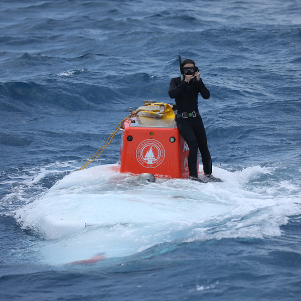 WHOI engineer Wren Sakai stands on the top of human occupied submersible Alvin as it prepares to descend. He is wearing a black wetsuit, snorkel gear, and. a weight belt.