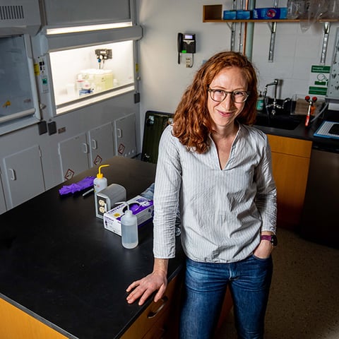 A woman with long hair and glasses leans against a table in a laboratory. She's smiling, with one hand in her pocket. Behind her is an illuminated bench fume hood.