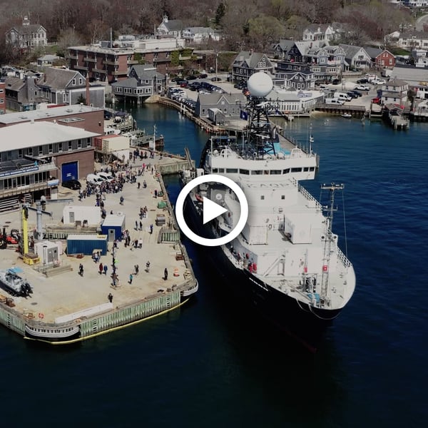 From a bird's eye view, WHOI's research vessel Atlantis (right) is parked in the water next to the Iselin Dock in Woods Hole. A group of people, small as ants, gather on the dock to welcome the ship and its crew.