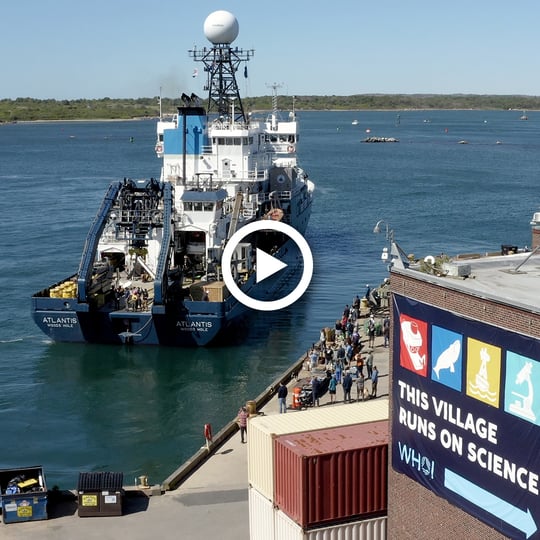 The research vessel Atlantis taxis out of Woods Hole harbor. A crowd sees the ship off from WHOI's Iselin Dock. In the foreground is a building with a banner that reads "this village runs on science."