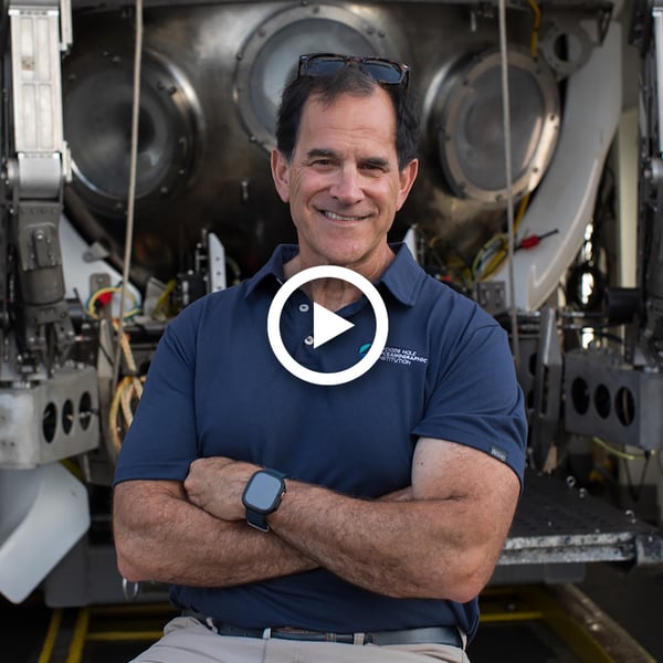 WHOI submersible pilot Bruce Strickrott smiles, arms crossed, in front of human-occupied vehicle Alvin. He's wearing a blue, Woods Hole Oceanographic Institution logo polo, a pair of sunglasses on his head, and an Apple watch.