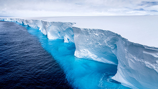 An aerial view showing deep water on the left and the jagged edge of an iceberg that extends into the horizon. Some of the iceberg is seen beneath the water's surface.