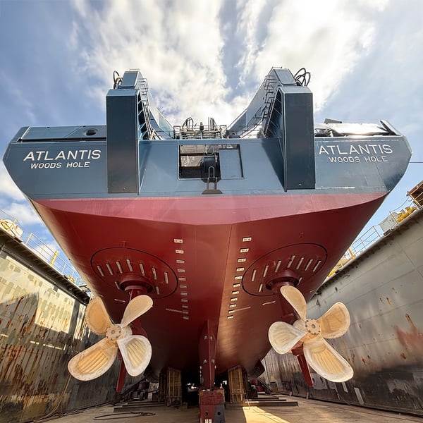 The underbelly of R/V Atlantis can be seen from below, a concrete wall on either side. Two large propellers are in view.