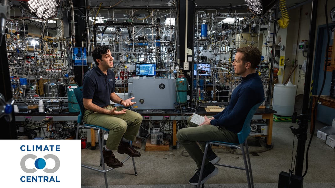 Two men in seats facing each other in a laboratory. The man on the left is wearing a polo with his hands out. The man on the right is holding a notepad and looking at the other man. Two studio lights are over their heads.