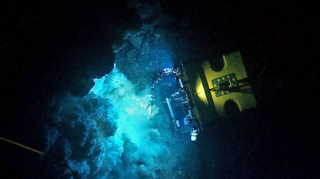 From above, an underwater robot with a mechanical arm shines its lights on a plume of smoke rising from the seafloor 