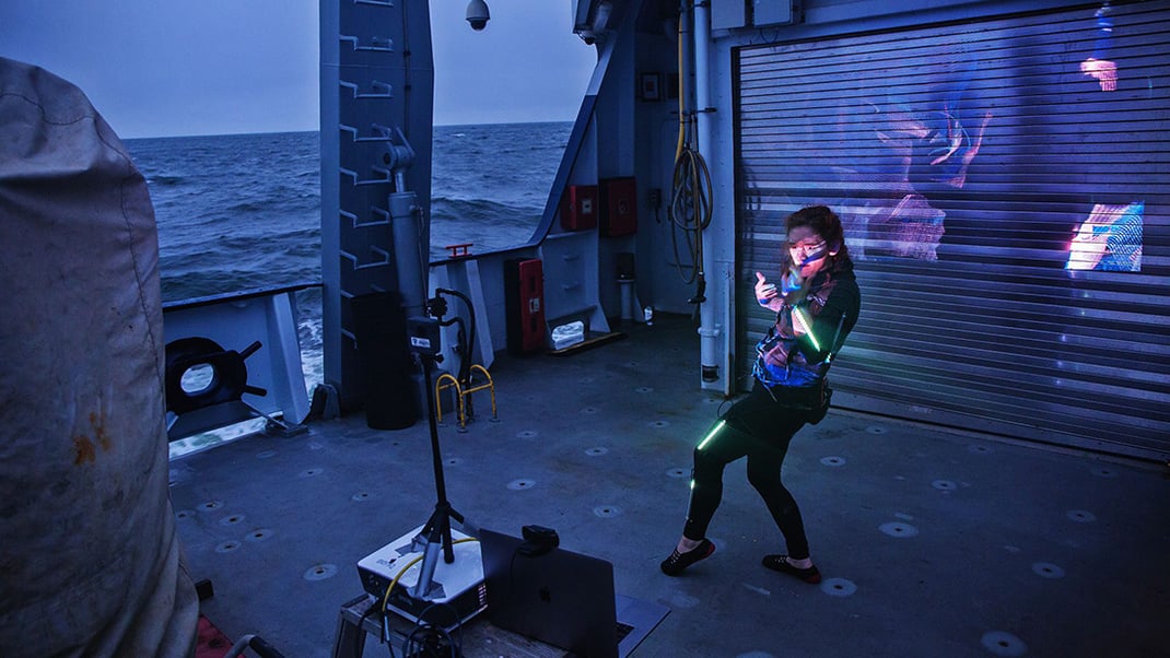 Artist Lori Hepner performs interpretative dance at dusk on the aft deck of a ship. She's wearing a black suit with LED light tubes affixed to her arms and legs. A projector is streaming lights in front of her.