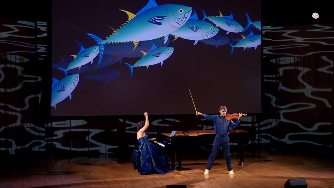 Two musicians perform on stage. (Left) a woman in a dress plays the piano lifting one hand up. (Right) A man with a button-up shirt tucked in plays the violin. Above them is a projector screen displaying cartoon images of yellowfin tuna.