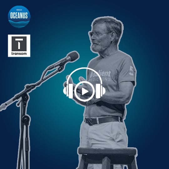 A bearded man wearing glasses and a T-shirt that says the word "Salient Predictions" on it faces left toward a microphone. His hands are held up while he speaks. 