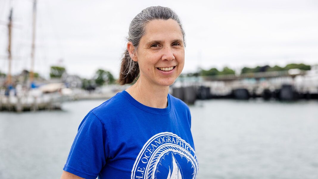 A portrait of WHOI Research Associate III Emily Peacock. She's wearing a blue T-shirt with a WHOI logo on it. Docks, a tall ship, and the water of Woods Hole harbor are behind her.
