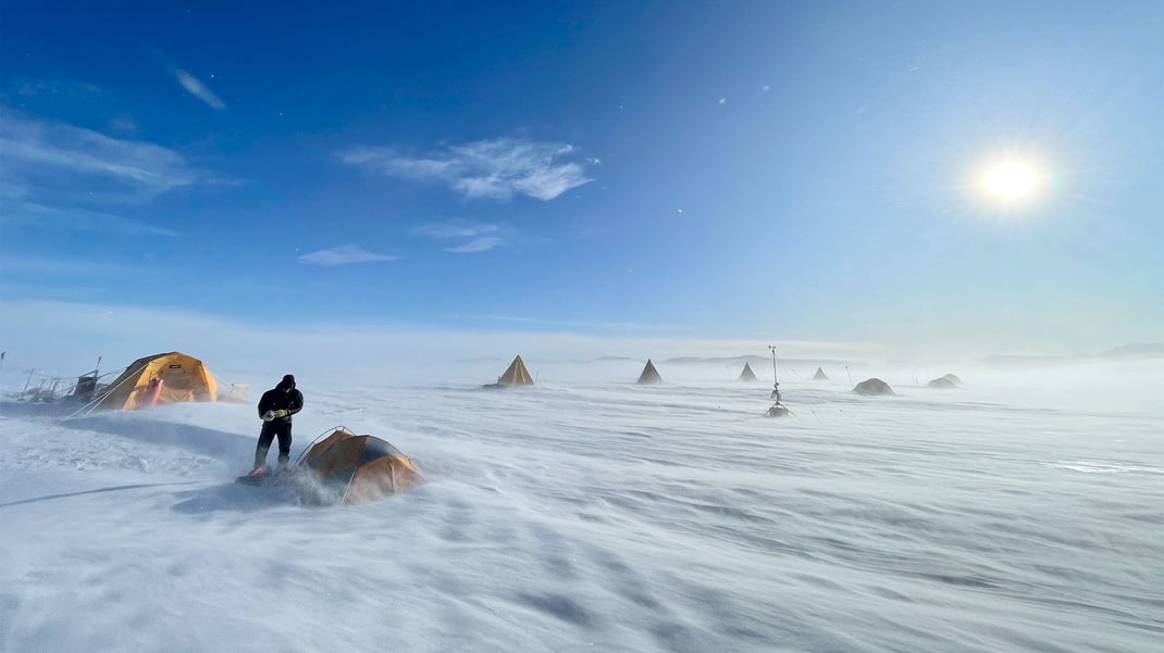 Eight tents on an open plane in Antarctic on a sunny day with clear skies. A lone figure appears outside one of the shelters as gusts of snow blow across the ground.